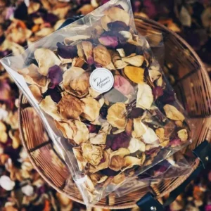 A clear plastic bag filled with natural mixed dried rose petals in shades of cream, pink, and burgundy, resting in a woven basket.