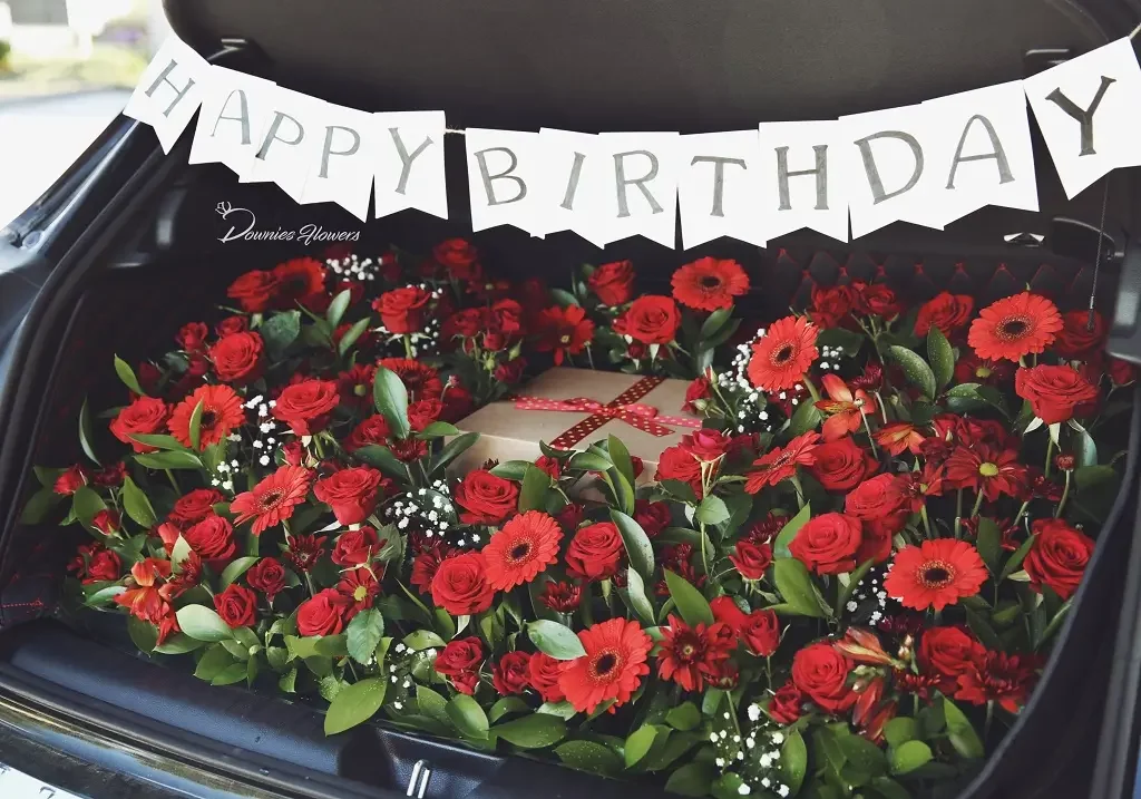 Car trunk filled with red roses and gerbera daisies, a birthday gift box, and a "HAPPY BIRTHDAY" banner in Christchurch. Same-day flower delivery available.